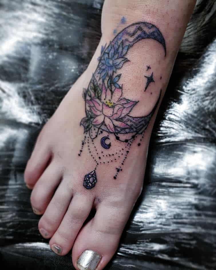 Elegant foot tattoo featuring a crescent moon adorned with blooming lotus flowers, delicate chandelier-style beadwork, a small crescent charm, and a faceted gemstone pendant