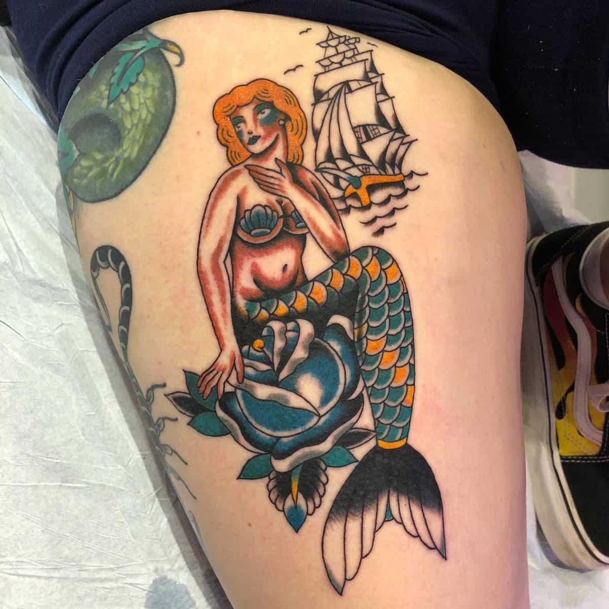 Colorful tattoo of a mermaid sitting on a flower with a sailing ship and sea creature in the background, on a thigh