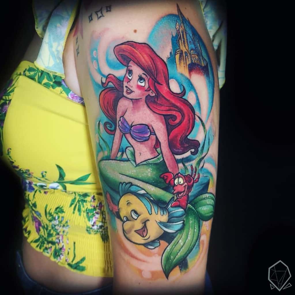 A colorful tattoo of Ariel with Flounder and Sebastian, surrounded by ocean swirls, a castle in the background, and vibrant details