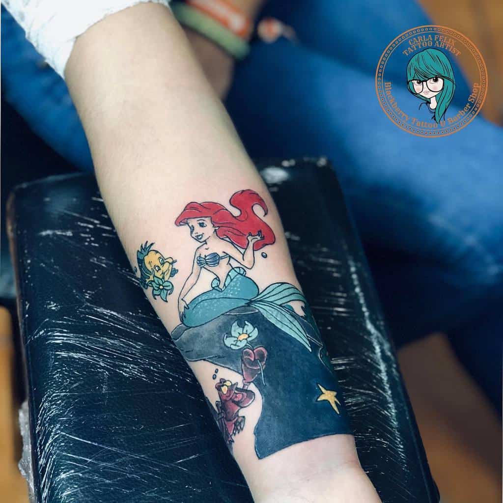 A charming tattoo of Ariel sitting on a rock with Flounder, Sebastian, and ocean details, featuring bold colors and playful elements