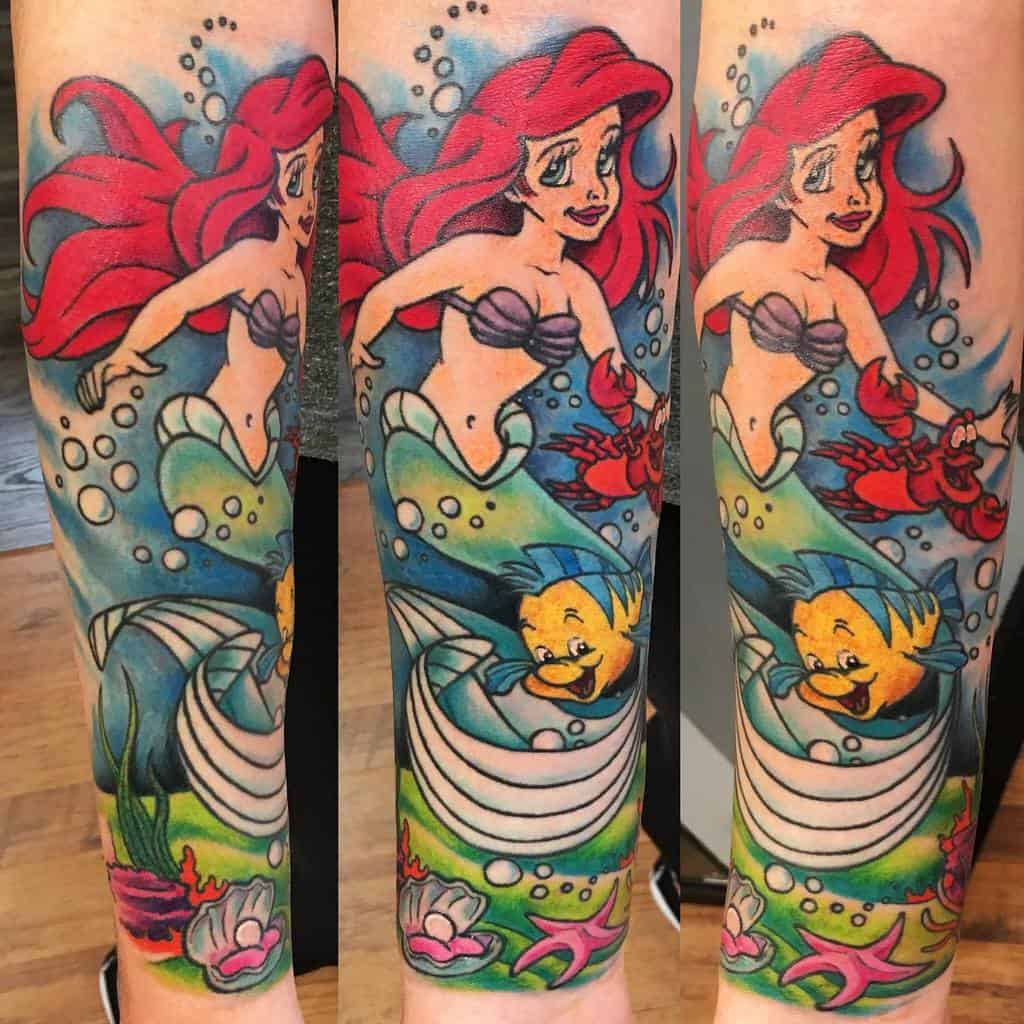 A vibrant full-arm tattoo of Ariel swimming with Flounder and Sebastian, surrounded by bubbles, sea plants, and colorful ocean scenery