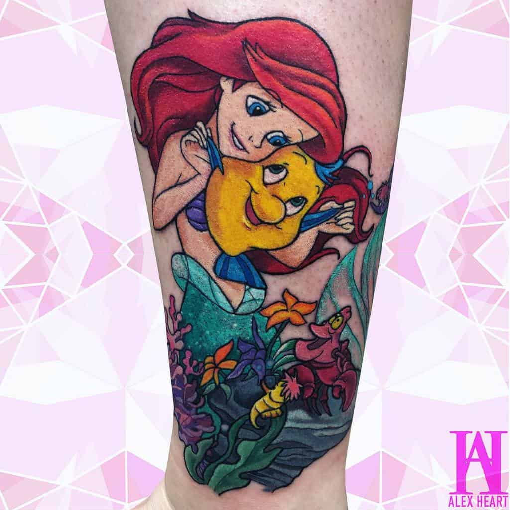 A vibrant tattoo of Ariel hugging Flounder, surrounded by colorful coral, flowers, and underwater details in a lively ocean scene