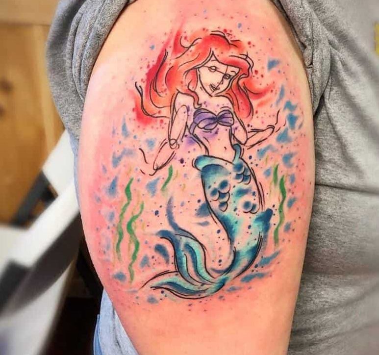 A whimsical watercolor-style tattoo of Ariel with vibrant red hair, a purple top, and a blue tail, surrounded by splashes of color and seaweed