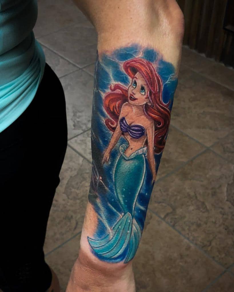 A detailed tattoo of Ariel with flowing red hair, a shimmering turquoise tail, and a magical underwater background in vibrant blue hues