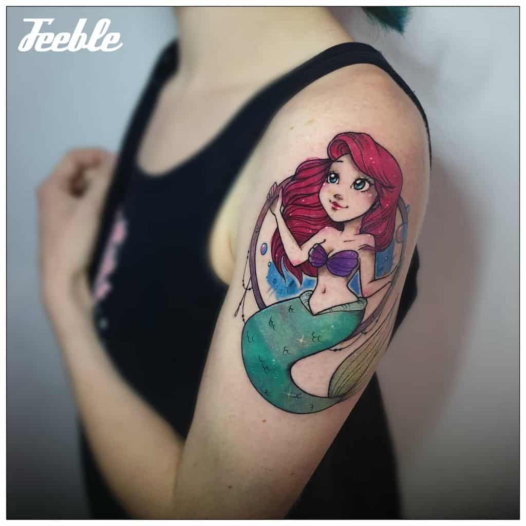 A whimsical tattoo of Ariel with big expressive eyes, vibrant red hair, and a green tail, set in a playful underwater scene on the upper arm