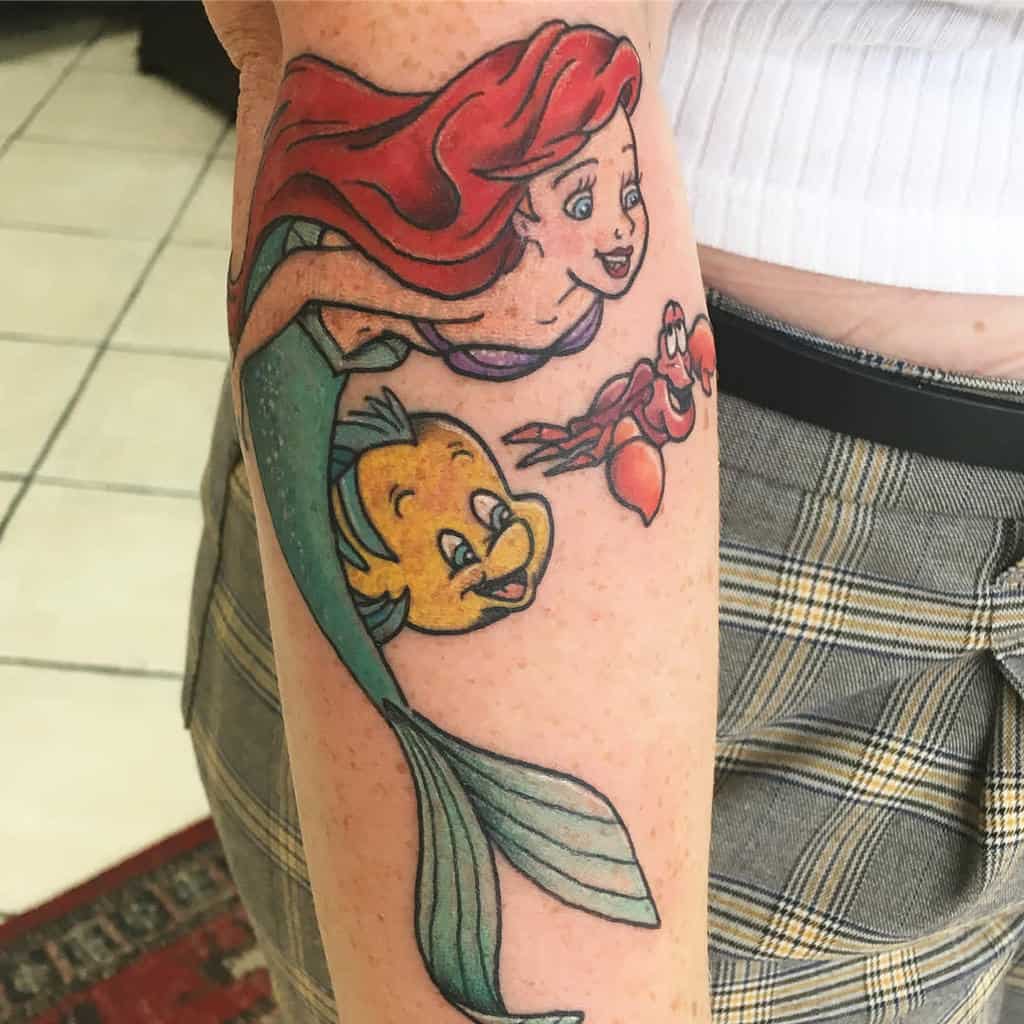A vibrant tattoo of Ariel swimming with Flounder and Sebastian on a forearm, capturing a joyful underwater moment