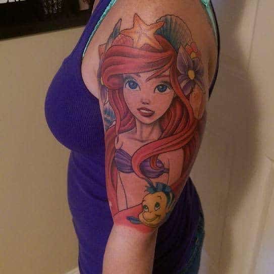 A detailed upper arm tattoo of Ariel from The Little Mermaid with vibrant red hair, surrounded by Flounder, seashells, and starfish accents