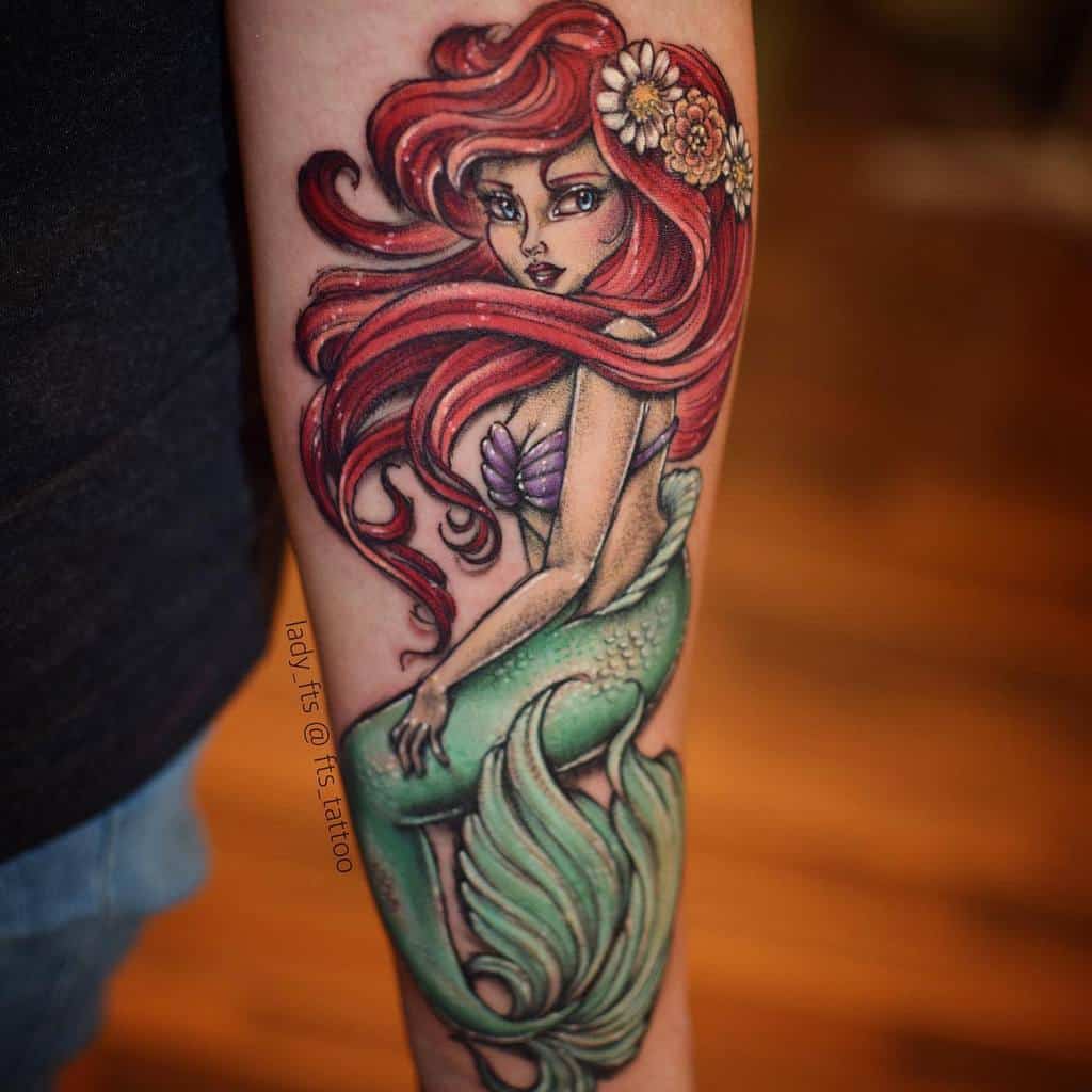 Intricate tattoo of Ariel from The Little Mermaid, featuring her signature flowing red hair, a seashell top, green mermaid tail, and floral accents in her hair, captured on a forearm with a vibrant and detailed design
