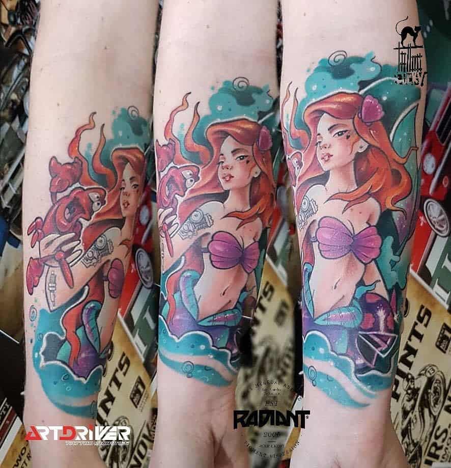 Tattoo of Ariel from The Little Mermaid on a forearm, featuring vibrant red hair, a purple seashell top, and Sebastian holding a fork