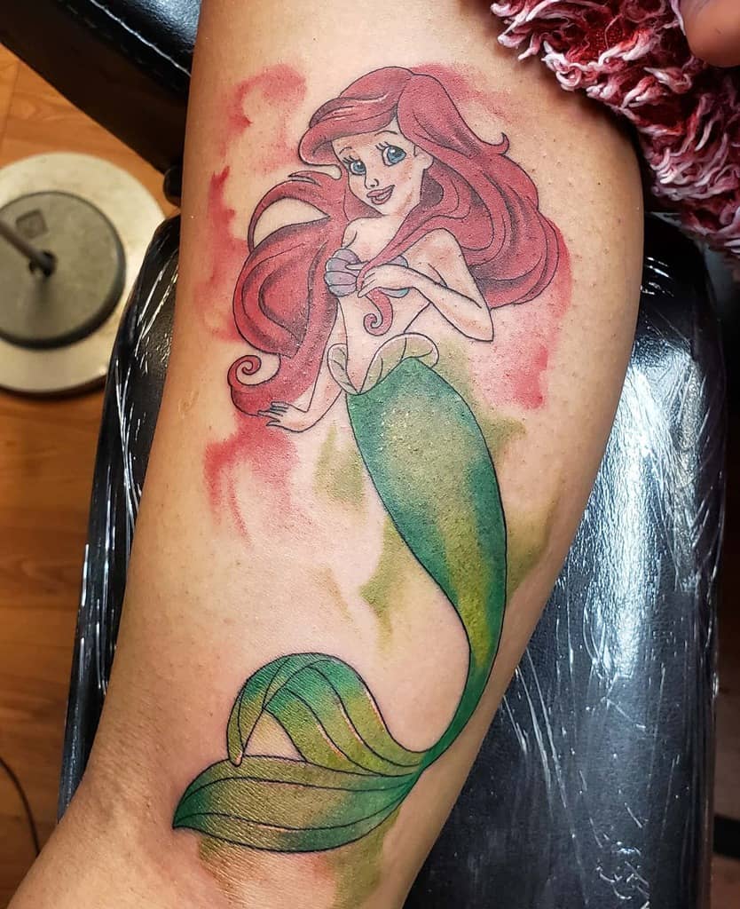 Beautiful tattoo of Ariel from The Little Mermaid with flowing red hair, green tail, and soft watercolor-style background on the calf
