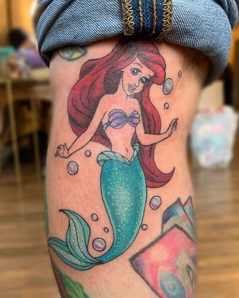 Playful Ariel tattoo with shimmering green tail, flowing red hair, and bubbles, showcasing a vibrant underwater vibe on the leg