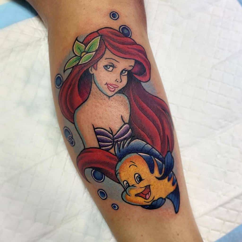 Bright and cheerful Ariel tattoo with a leafy hair accessory, flowing red hair, and Flounder surrounded by bubbles on the leg