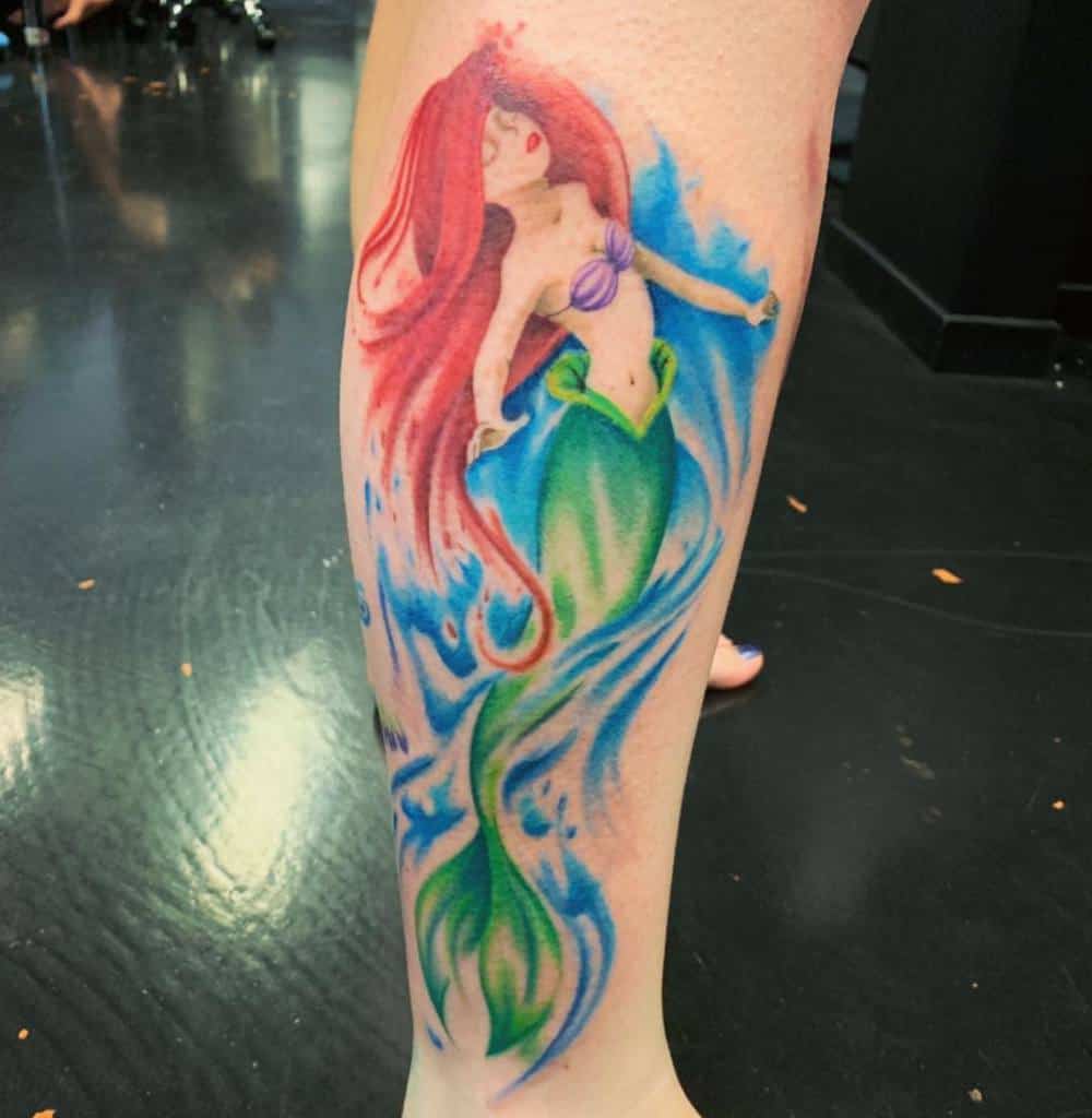 Whimsical Ariel tattoo with flowing red hair, vibrant green tail, and a dreamy watercolor-style blue background on the leg