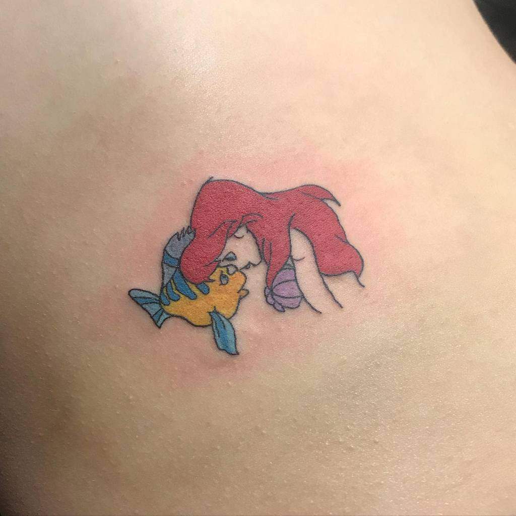Delicate and minimal Ariel tattoo sharing a sweet moment with Flounder, featuring soft colors and a small, simple design