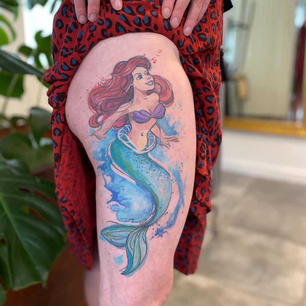 Vibrant watercolor thigh tattoo showcasing Ariel in motion, surrounded by splashes of blue and pink hues for a dreamy aquatic effect