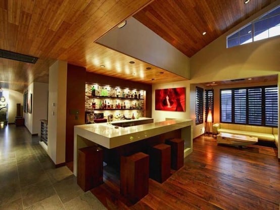 Modern home bar with wooden ceiling, backlit shelves, and a sleek white counter with four stools in a cozy setting