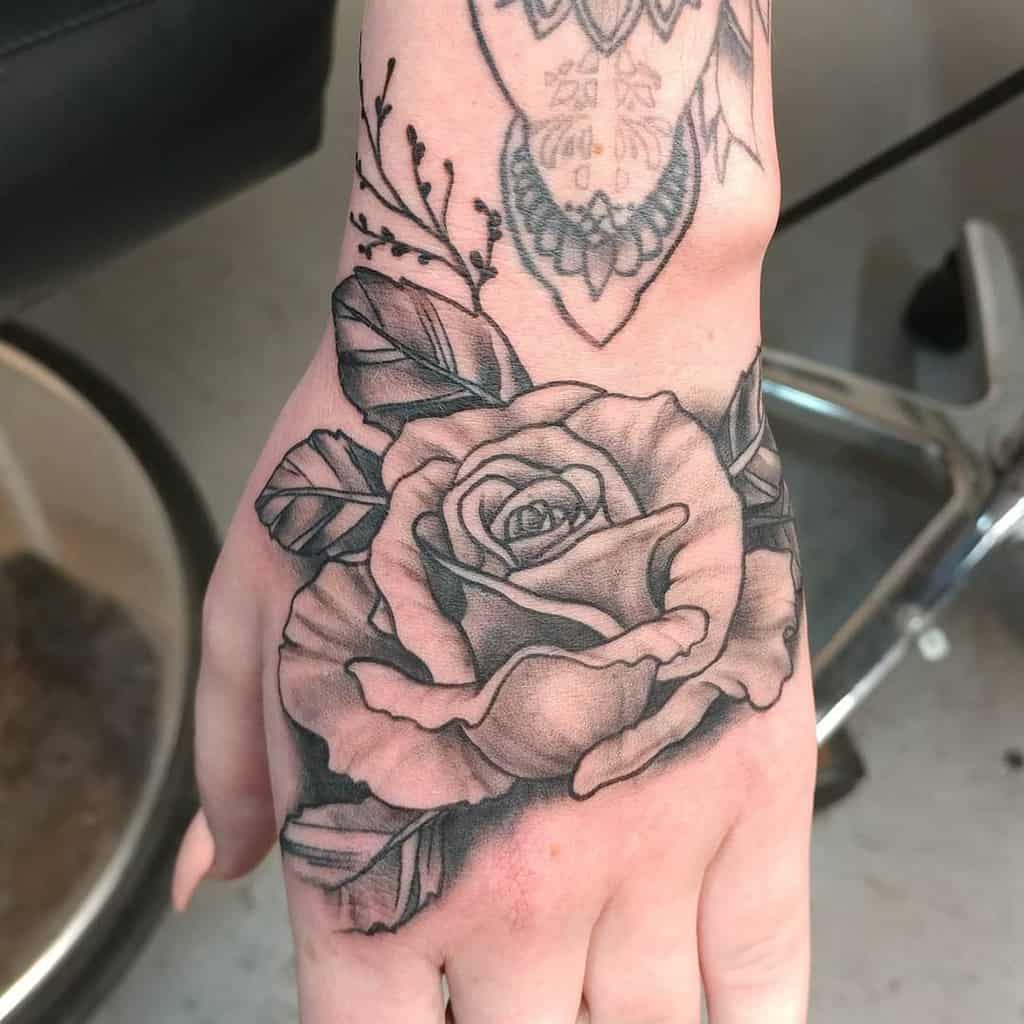 Hand tattoo featuring a realistic black and grey rose with detailed shading and leaves, complemented by ornamental wrist tattoo elements