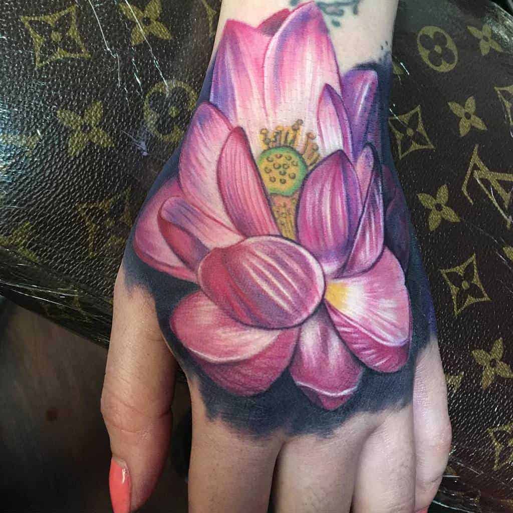Tattoo of a pink lotus flower on a person's hand against a patterned background