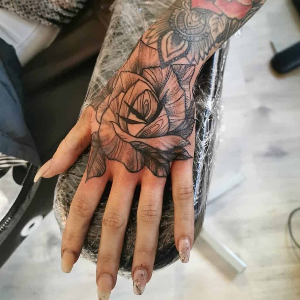 Hand tattoo featuring a bold black and grey rose design, with intricate shading and ornamental details