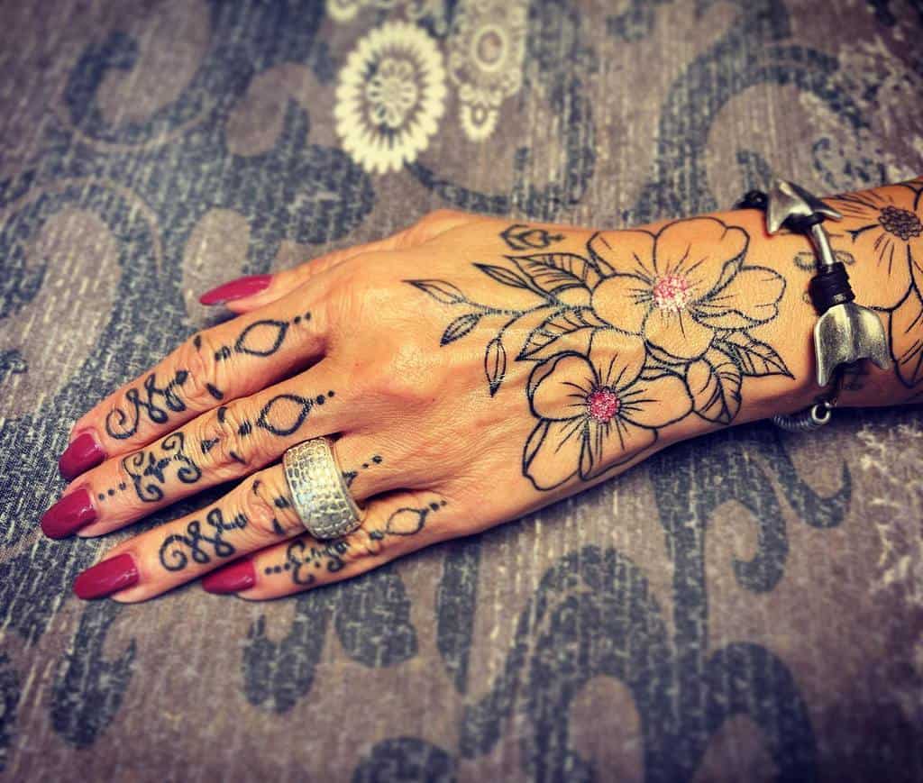 Hand and finger tattoos featuring floral designs with pink-centered blossoms, ornamental script, and patterns