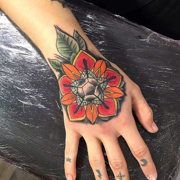 A hand with a colorful geometric flower tattoo featuring red and orange petals, green leaves, and a hexagonal center, with star, lightning, crus wit four dots and crescent moon