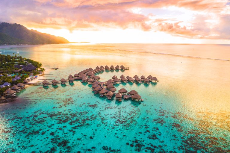 Aerial view of overwater bungalows on turquoise water at sunset, lush mountains and colorful sky in the background, luxury travel