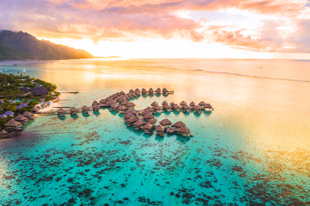 Aerial view of overwater bungalows on turquoise water at sunset, lush mountains and colorful sky in the background, luxury travel