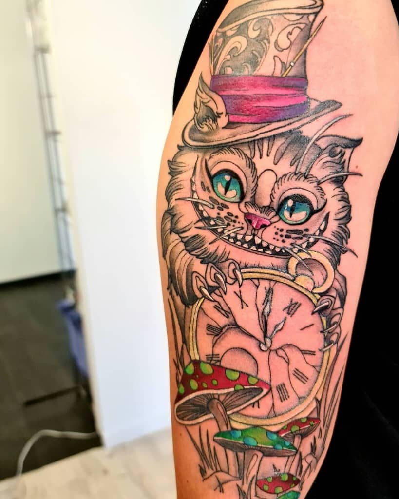 A detailed tattoo of the Cheshire Cat wearing a Mad Hatter hat, holding a cracked clock, with colorful mushrooms at the bottom