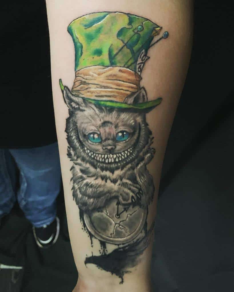 A vibrant Cheshire Cat tattoo wearing a green Mad Hatter hat, holding a cracked clock, with a wide grin and glowing blue eyes