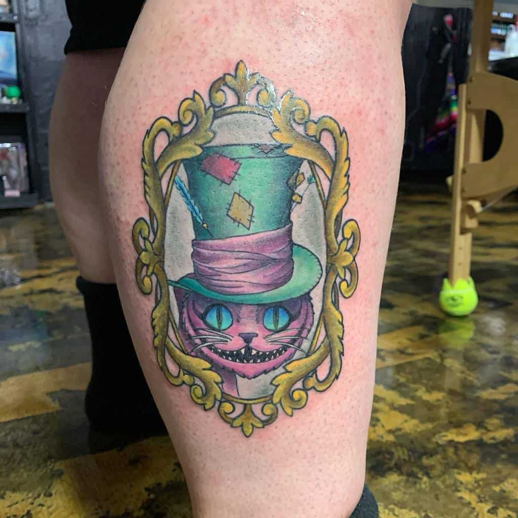 A colorful Cheshire Cat tattoo wearing a patched-up Mad Hatter hat, framed in a golden ornate border, with glowing blue eyes and a mischievous grin