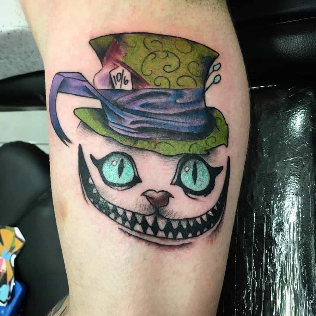 A whimsical Cheshire Cat tattoo with glowing blue eyes and a wide grin, wearing a green Mad Hatter hat with the "10/6" label
