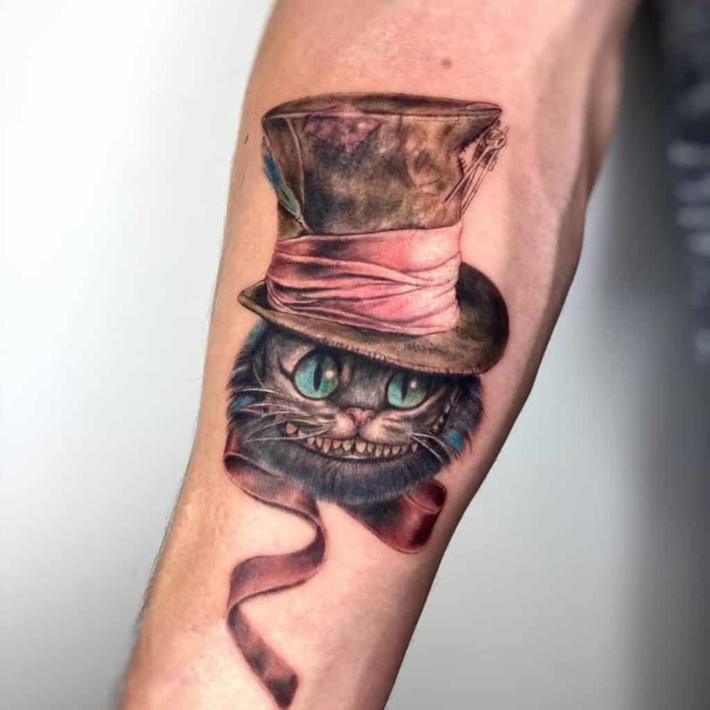 A tattoo of the Cheshire Cat wearing a tattered Mad Hatter hat with a pink ribbon, featuring the cat's mischievous grin and glowing green eyes, creating a whimsical yet slightly eerie vibe