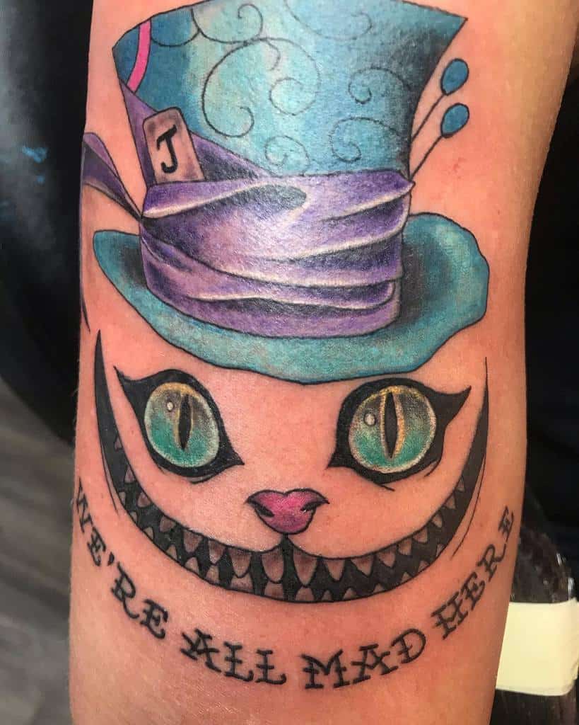 Cheshire Cat tattoo featuring a Mad Hatter hat and "We're All Mad Here" text, embracing Wonderland's charm