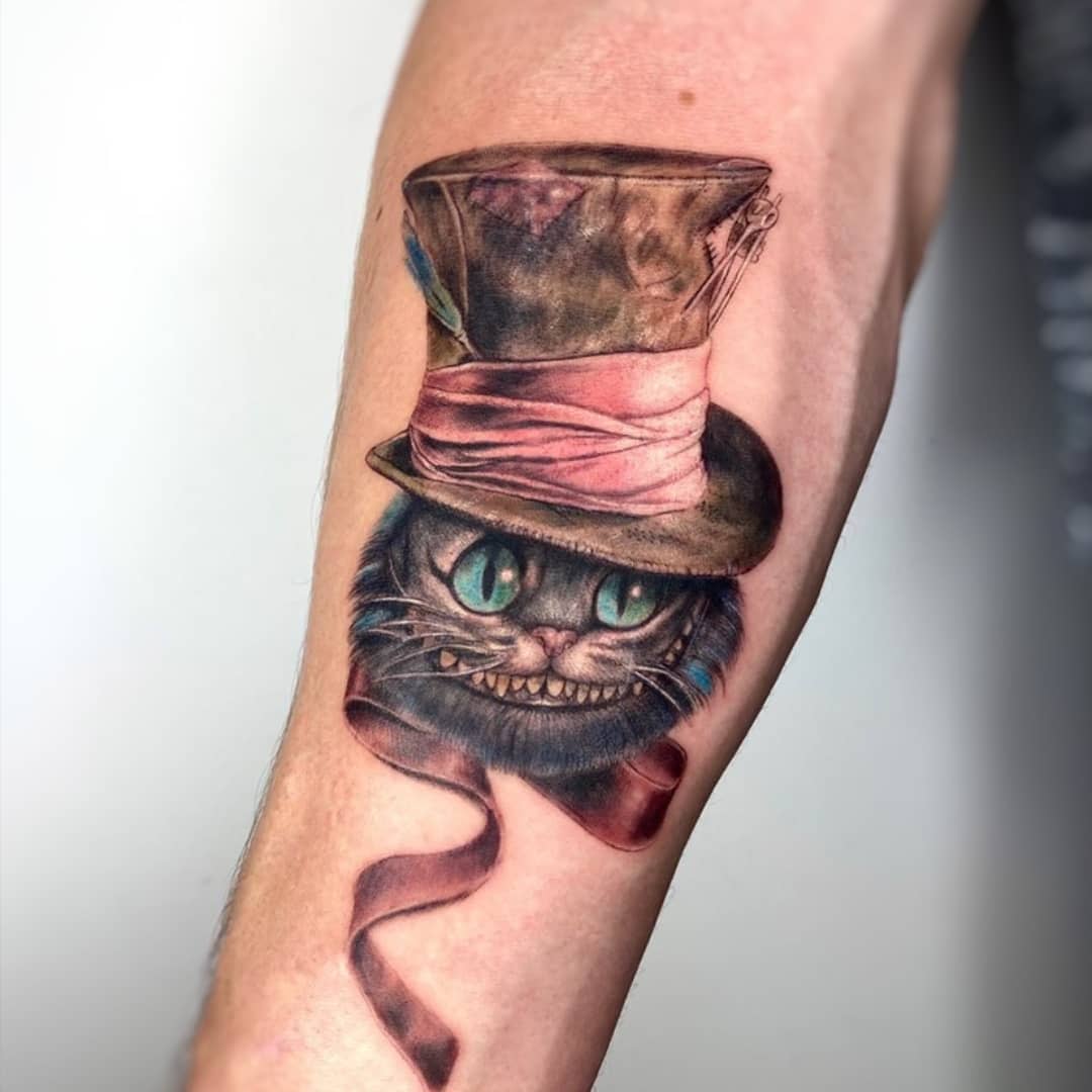 Realistic tattoo of the Cheshire Cat wearing a worn Mad Hatter hat, with a mischievous grin and ribbon