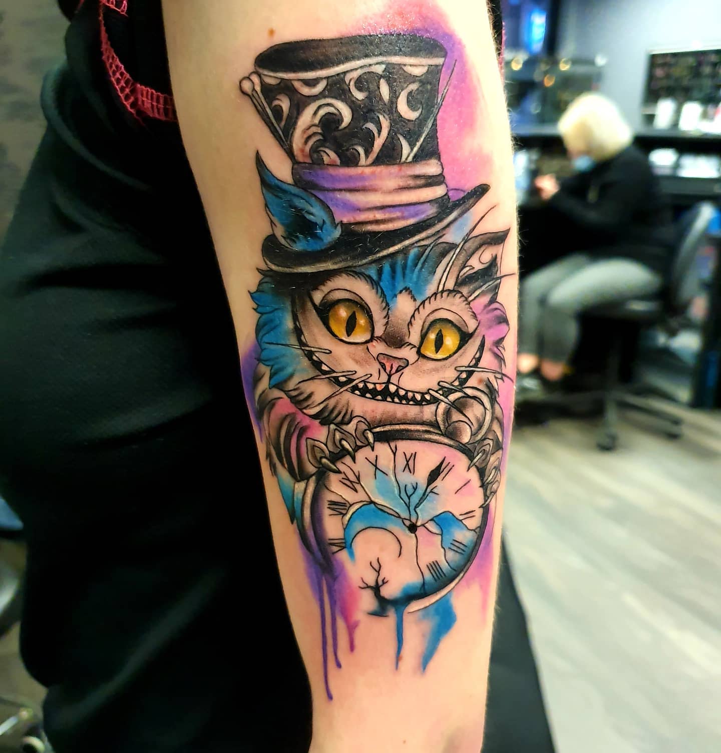 Watercolor tattoo of the Cheshire Cat wearing a Mad Hatter hat, holding a cracked pocket watch, with vibrant purple and blue splashes