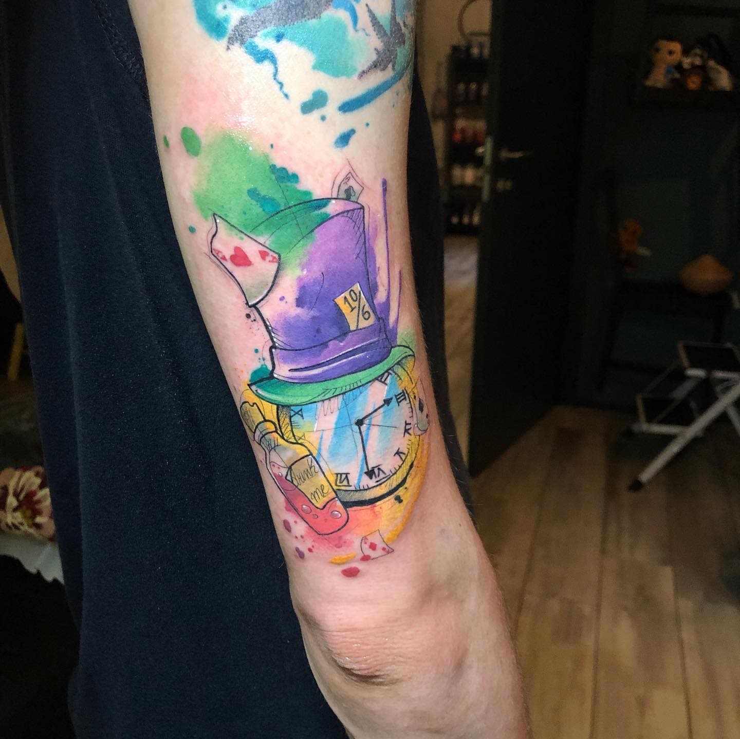 Watercolor tattoo of the Mad Hatter’s hat with a pocket watch, playing cards, and a 'Drink Me' bottle, accented with splashes of vibrant colors