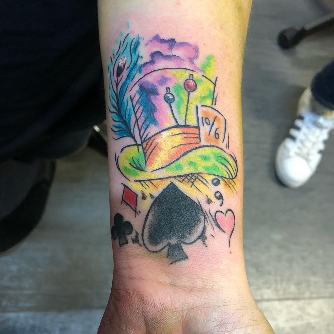 Watercolor tattoo of the Mad Hatter’s hat with a peacock feather, playing card symbols, and vibrant splashes of color