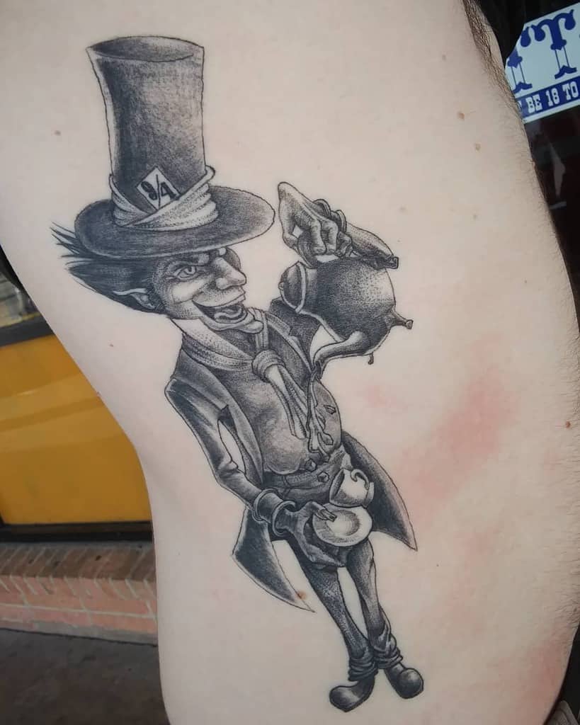 Black and gray tattoo of the Mad Hatter from Alice in Wonderland, holding a teapot and pouring it in a cup, with his signature '9/4' hat