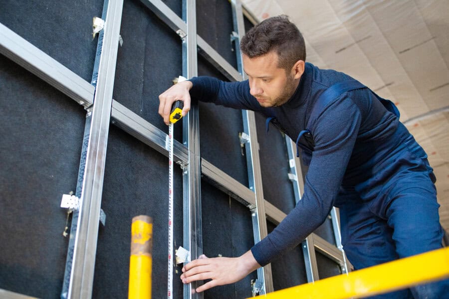 A man measures a metal frame with a tape measure, working carefully on a construction or renovation project indoors