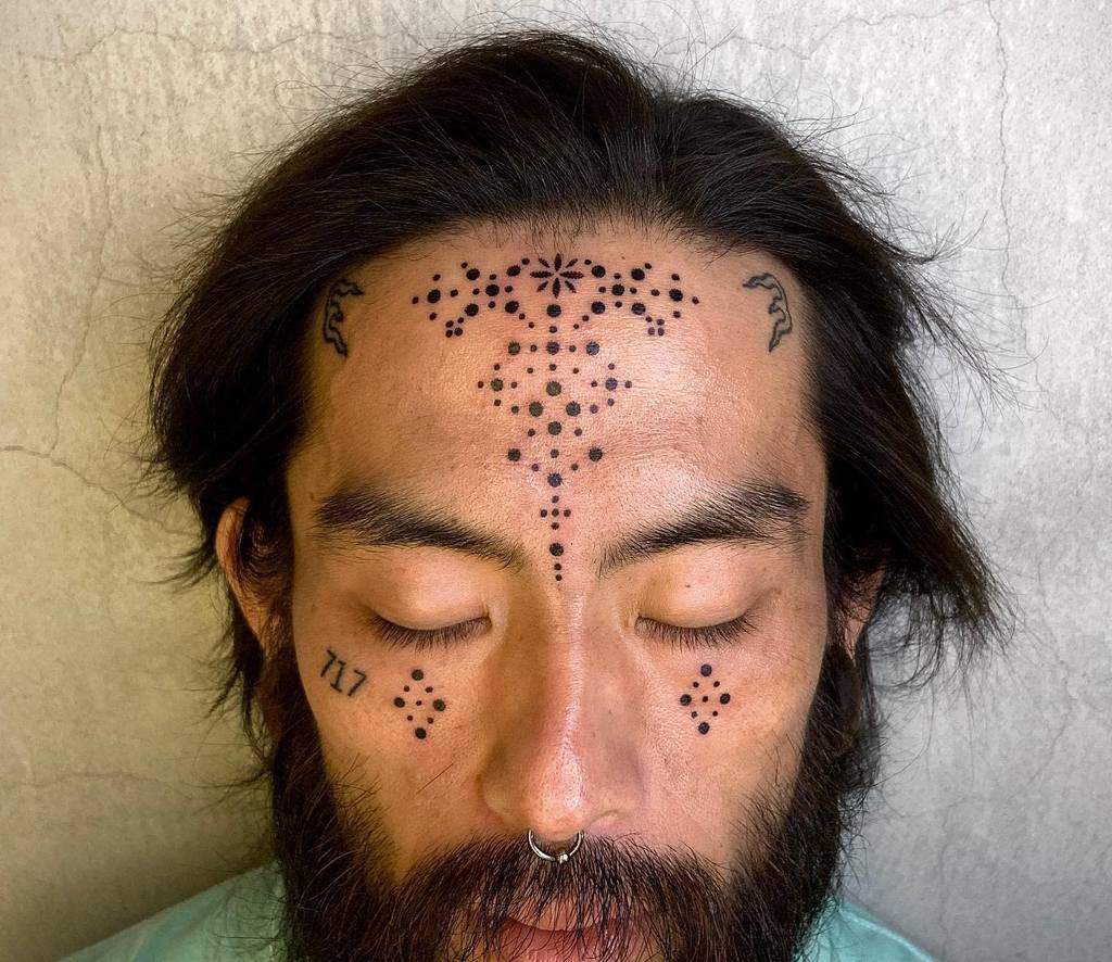 A man with tribal-style face tattoos featuring symmetrical dot patterns on his forehead, cheeks, and under his eyes, including the number "717" under the left eye