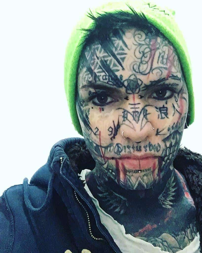 A person with highly detailed face tattoos, covering nearly their entire face with geometric, script, and symbolic designs