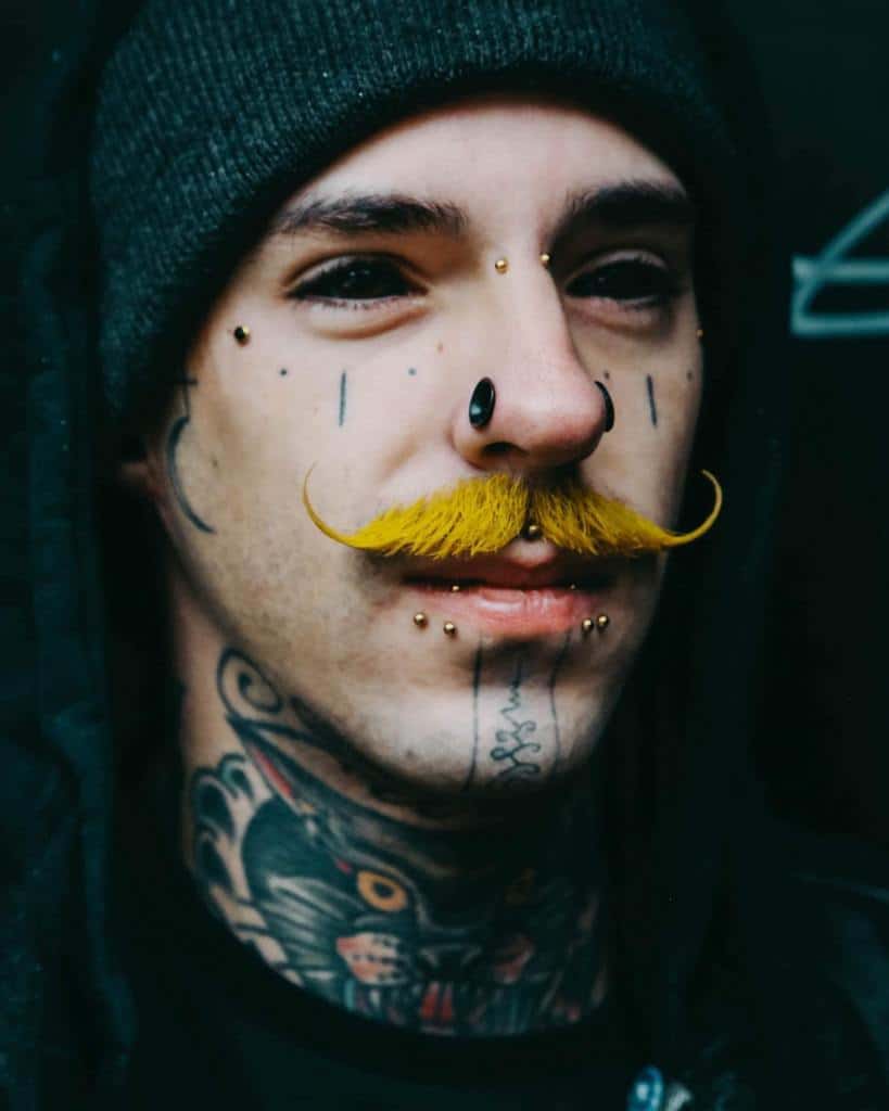 Man with multiple piercings and bold tattoos, including teardrop symbols near the eyes and a bold neck tattoo
