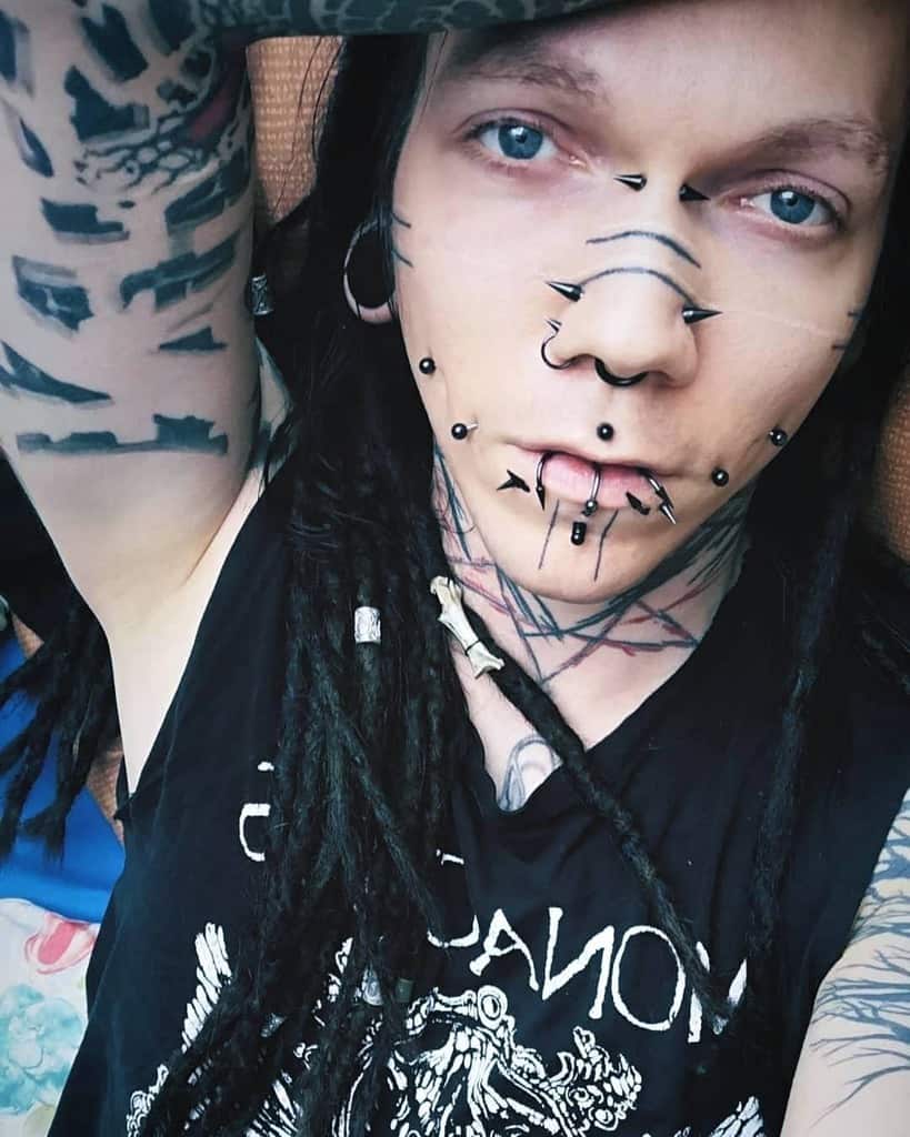 Man with multiple piercings and geometric face tattoos, including symmetrical lines and bold designs across the forehead and chin