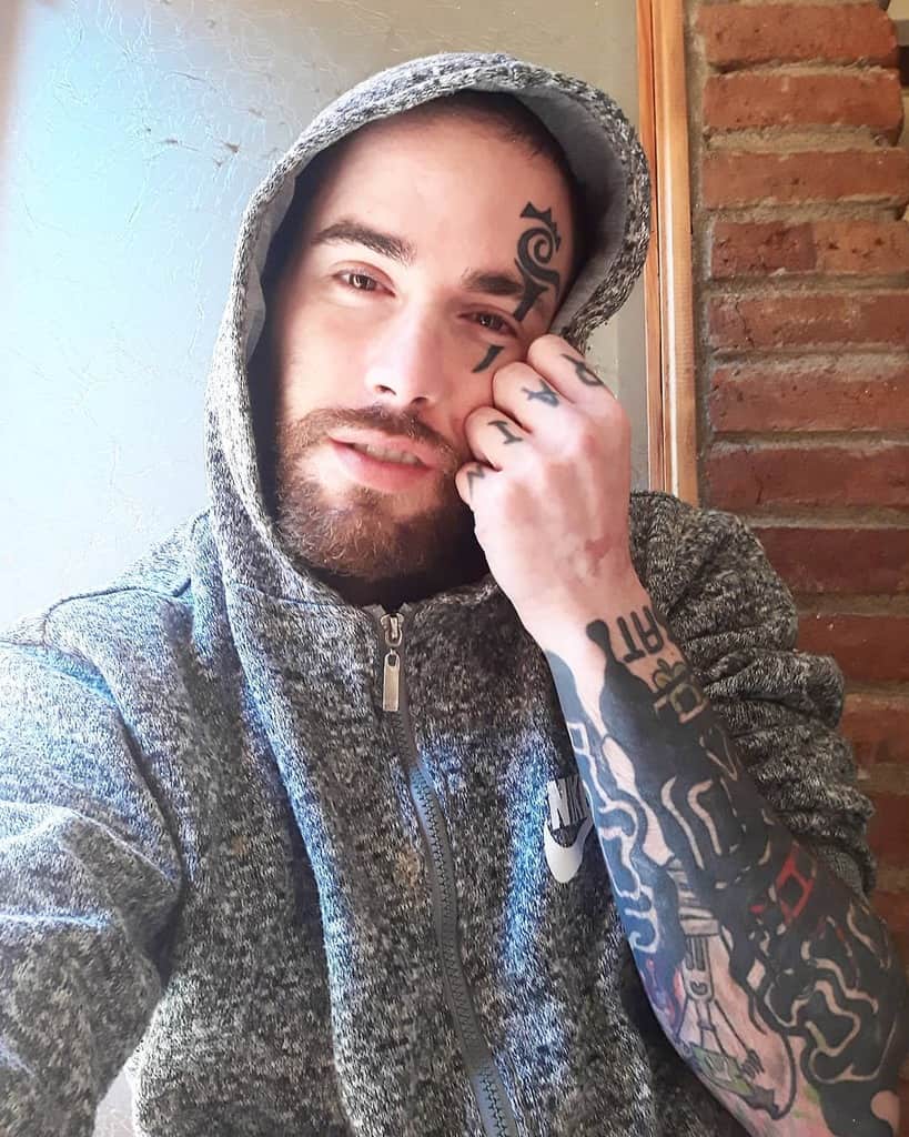 Man with a bold tribal-style tattoo on his temple and hand, wearing a hoodie with intricate sleeve tattoos on his arm