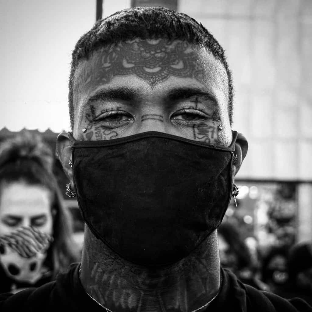 Black and white portrait of a man with intricate forehead and cheek tattoos, wearing a face mask and earrings