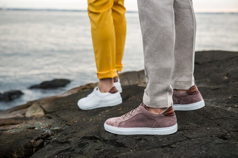 Two people stand on rocks near water, wearing vibrant pants and stylish men's shoes in modern white and mauve shoe designs