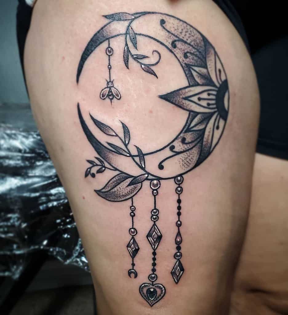 Tattoo of a crescent moon with intricate floral patterns and delicate hanging ornaments on a person's leg