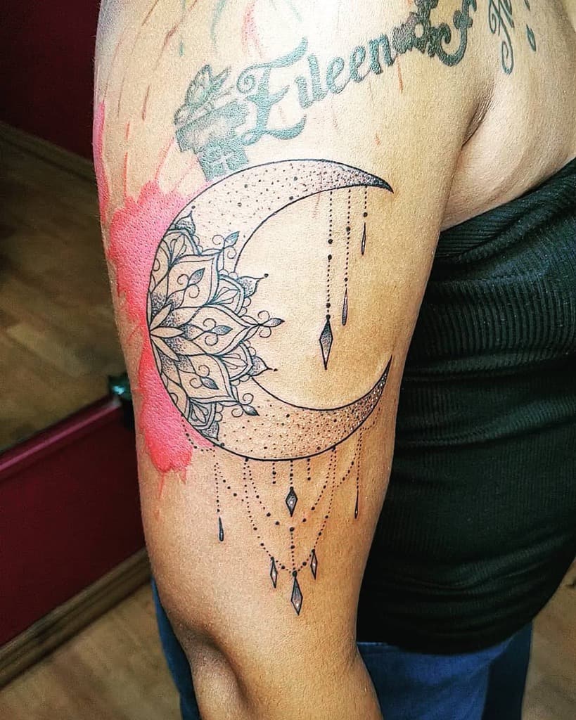 Tattoo of an intricate crescent moon design with dangling ornaments on a person's upper arm