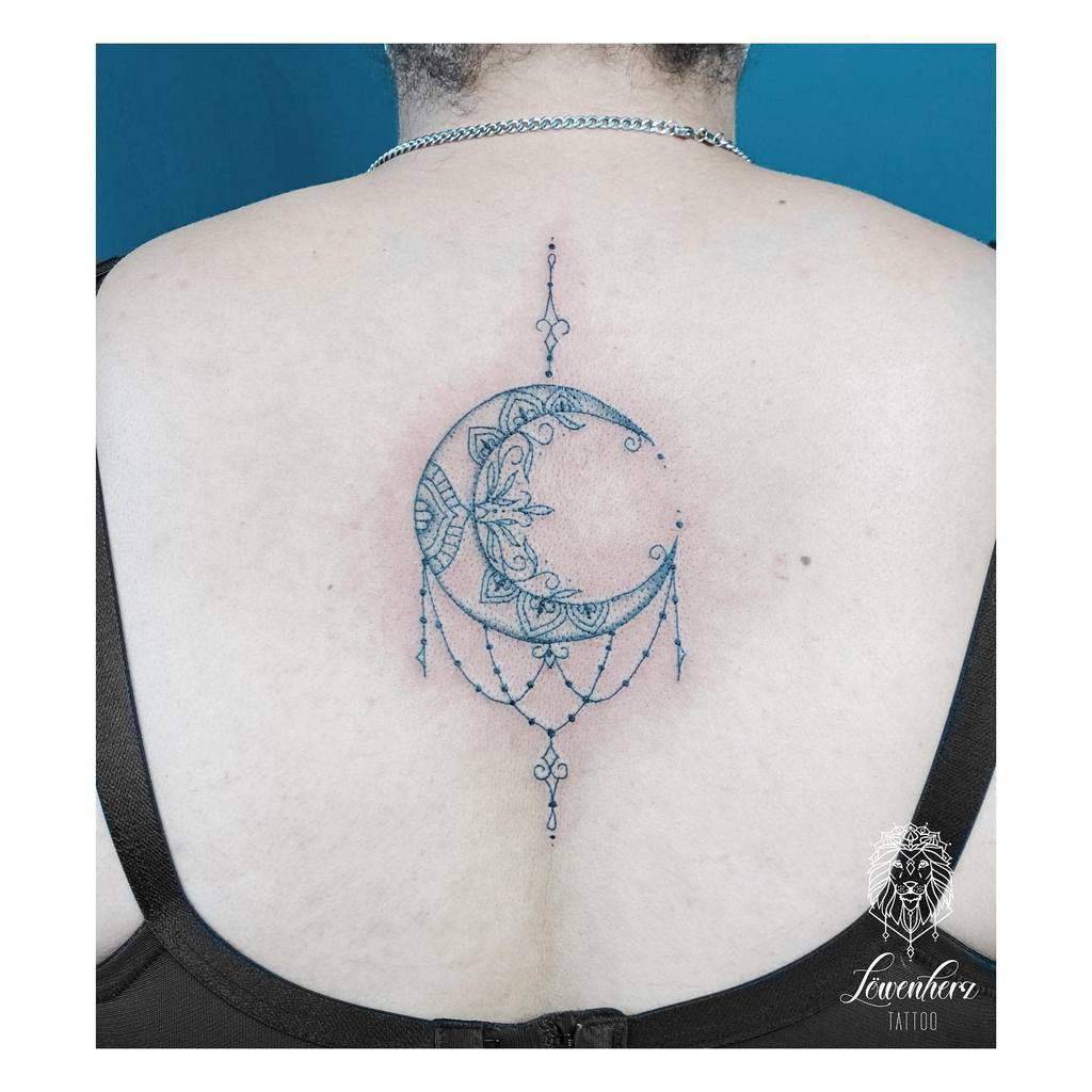 Intricate crescent moon tattoo with decorative patterns and dangling elements inked on a person’s back