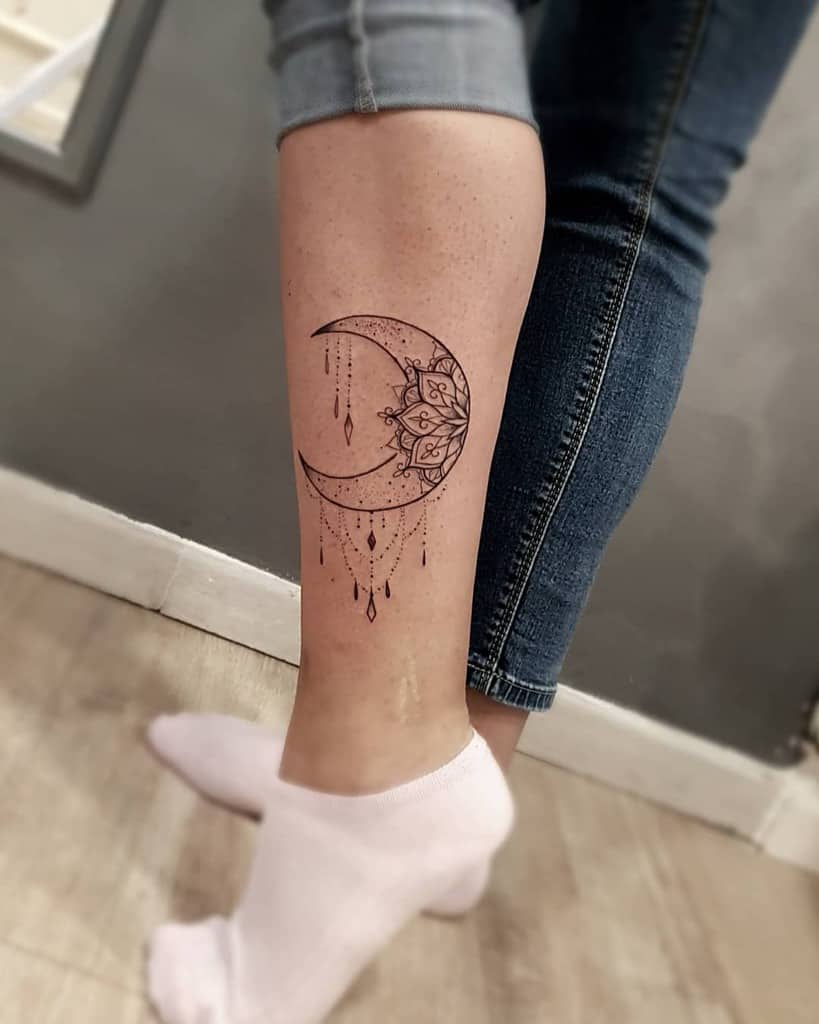 Tattoo of a crescent moon with intricate designs on the back of a lower leg